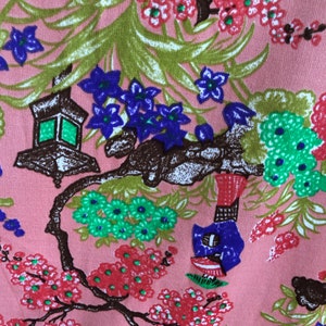 May include: Pink fabric with a floral pattern featuring a brown tree branch with green and blue flowers, a lantern, and a blue bird. The background is a light pink color.