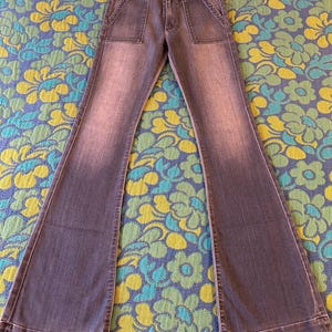 May include: Faded blue denim bell-bottom jeans with two front pockets. The jeans have a high waist and a button and zipper closure. The legs flare out dramatically from the knee down. The jeans are laid flat on a floral patterned surface.