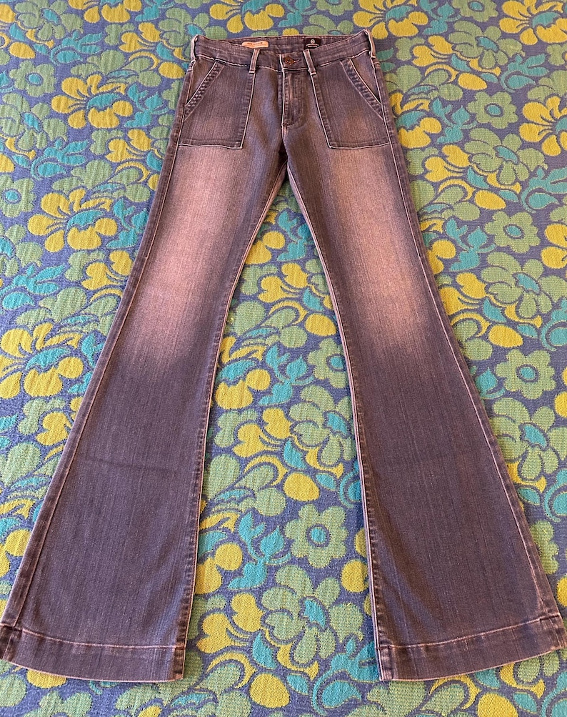 May include: Faded blue denim bell-bottom jeans with two front pockets. The jeans have a high waist and a button and zipper closure. The legs flare out dramatically from the knee down. The jeans are laid flat on a floral patterned surface.