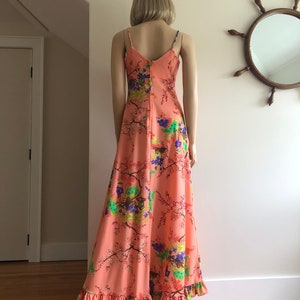 May include: A long, peach-colored maxi dress with a floral print featuring pink, green, blue, and purple flowers. The dress has thin straps and a ruffled hem.