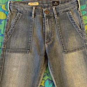 May include: Light blue denim jeans with large front pockets and a button closure. The jeans have a faded wash and a classic five-pocket design. The brand name "AG Goldie" is visible on the label.