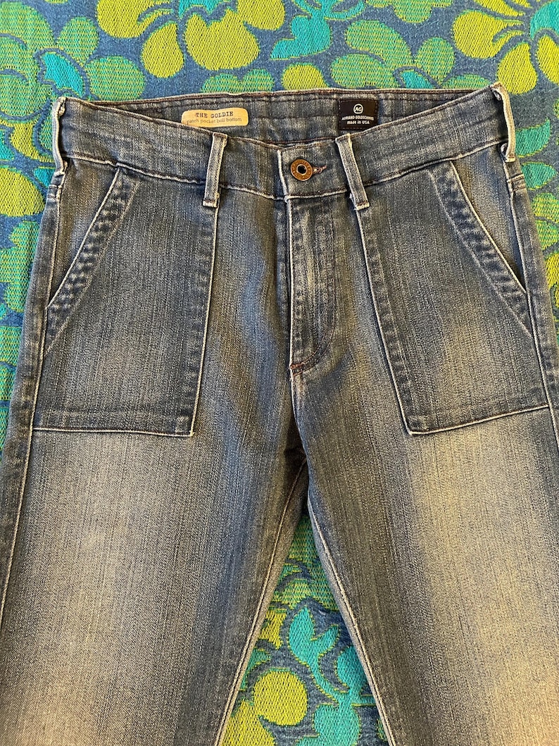 May include: Light blue denim jeans with large front pockets and a button closure. The jeans have a faded wash and a classic five-pocket design. The brand name "AG Goldie" is visible on the label.