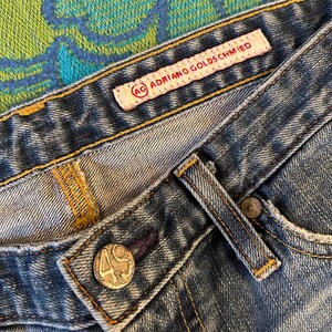 May include: Close-up of blue denim jeans with visible stitching and a button. The jeans feature a label that reads "AG Adriano Goldschmied". The button has the number "49" on it.