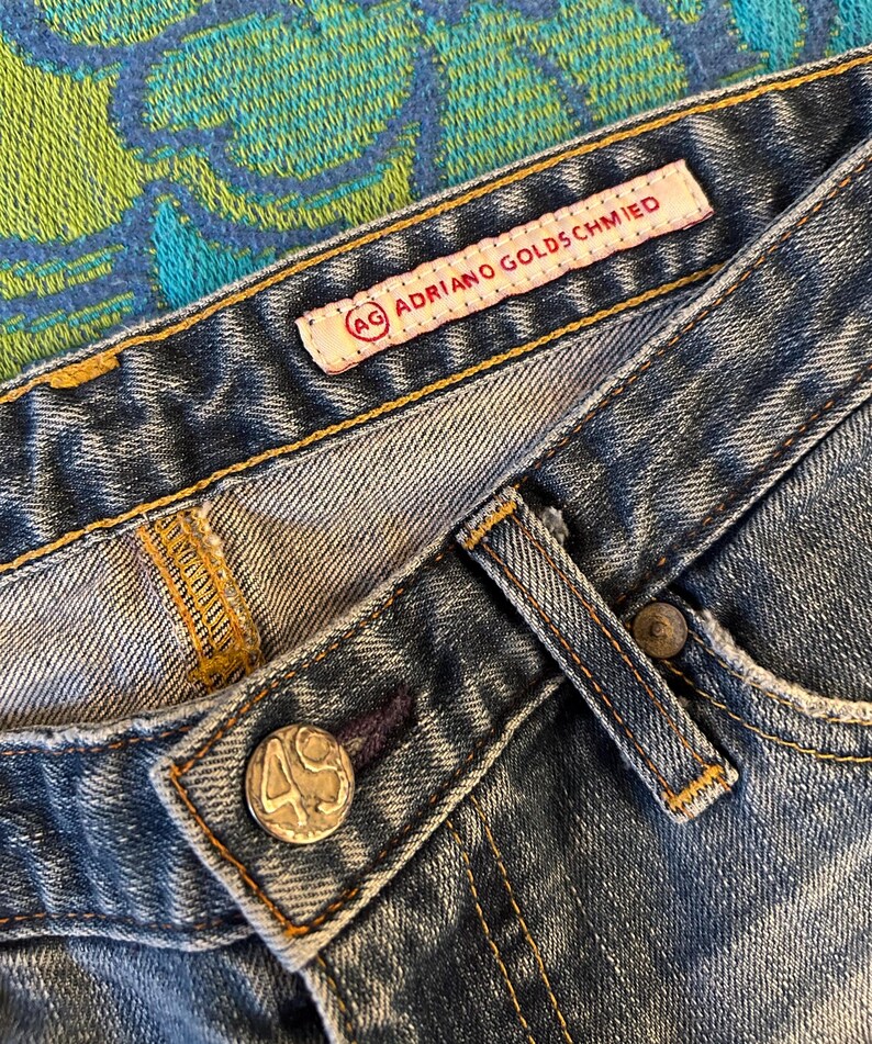May include: Close-up of blue denim jeans with visible stitching and a button. The jeans feature a label that reads "AG Adriano Goldschmied". The button has the number "49" on it.
