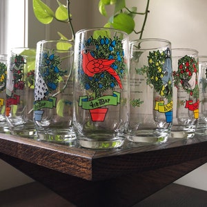 May include: Set of 10 clear glass tumblers with colorful illustrations of Christmas trees and birds. Each glass has a different bird and a banner with the number of the day of Christmas. The glasses are arranged in a row on a wooden surface.