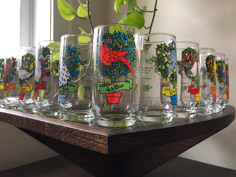 May include: Set of 10 clear glass tumblers with colorful illustrations of Christmas trees and birds. Each glass has a different bird and a banner with the number of the day of Christmas. The glasses are arranged in a row on a wooden surface.