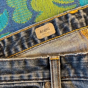 May include: Close-up of blue denim jeans with a light wash. The jeans have a brown label that reads "Angel" and "boot cut". The jeans have copper-coloured rivets and stitching.