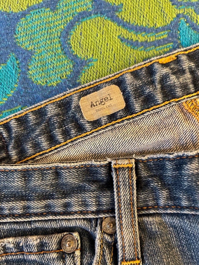 May include: Close-up of blue denim jeans with a light wash. The jeans have a brown label that reads "Angel" and "boot cut". The jeans have copper-coloured rivets and stitching.