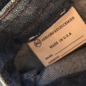 May include: Close-up of a denim garment with a label. The label reads "ADRIANO GOLDSCHMIED" and "MADE IN U.S.A." with space for a name and address. The denim is a medium blue with visible stitching.
