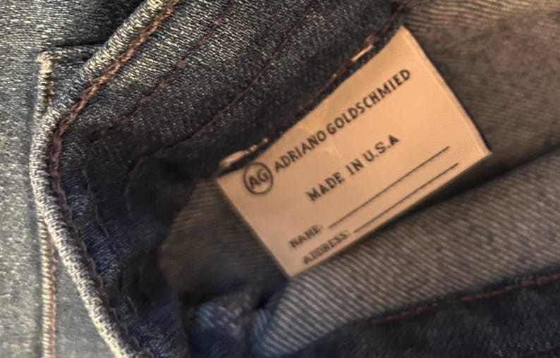 May include: Close-up of a denim garment with a label. The label reads "ADRIANO GOLDSCHMIED" and "MADE IN U.S.A." with space for a name and address. The denim is a medium blue with visible stitching.