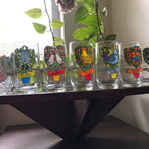 May include: Set of 12 glass tumblers with colorful illustrations of Christmas trees and the days of Christmas. Each glass features a different tree with a unique design and the corresponding day of Christmas. The glasses are arranged in a row on a wooden shelf.