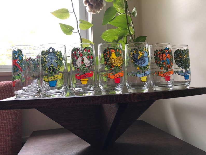 May include: Set of 12 glass tumblers with colorful illustrations of Christmas trees and the days of Christmas. Each glass features a different tree with a unique design and the corresponding day of Christmas. The glasses are arranged in a row on a wooden shelf.