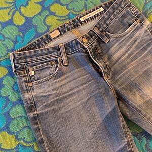 May include: Close-up of blue denim jeans with a faded wash. The jeans have a classic five-pocket design, a button and zip closure, and belt loops. The brand name is visible on the waistband.