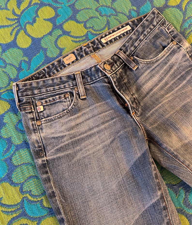 May include: Close-up of blue denim jeans with a faded wash. The jeans have a classic five-pocket design, a button and zip closure, and belt loops. The brand name is visible on the waistband.
