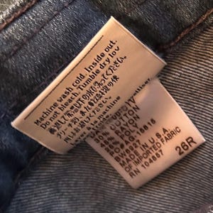 May include: A white fabric garment care tag sewn into dark blue denim. The tag features black text with washing instructions in English, Japanese, and French, along with a material composition of 30% cotton, 45% rayon, 25% poly, and the size 26R.