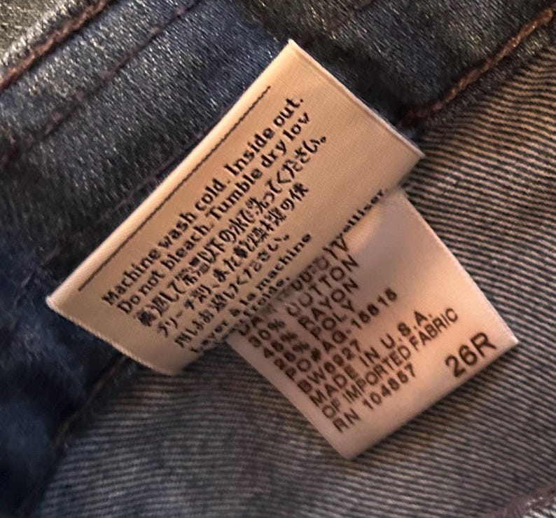 May include: A white fabric garment care tag sewn into dark blue denim. The tag features black text with washing instructions in English, Japanese, and French, along with a material composition of 30% cotton, 45% rayon, 25% poly, and the size 26R.