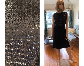 May include: A black sleeveless dress with a sequined bodice and a black skirt. The dress is on a mannequin in a room with a wooden floor.