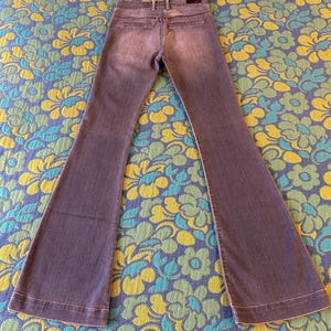 May include: A pair of gray denim bell-bottom jeans. The jeans have a classic five-pocket design, a button and zipper closure, and a flared leg. The jeans are laid flat on a floral patterned surface.