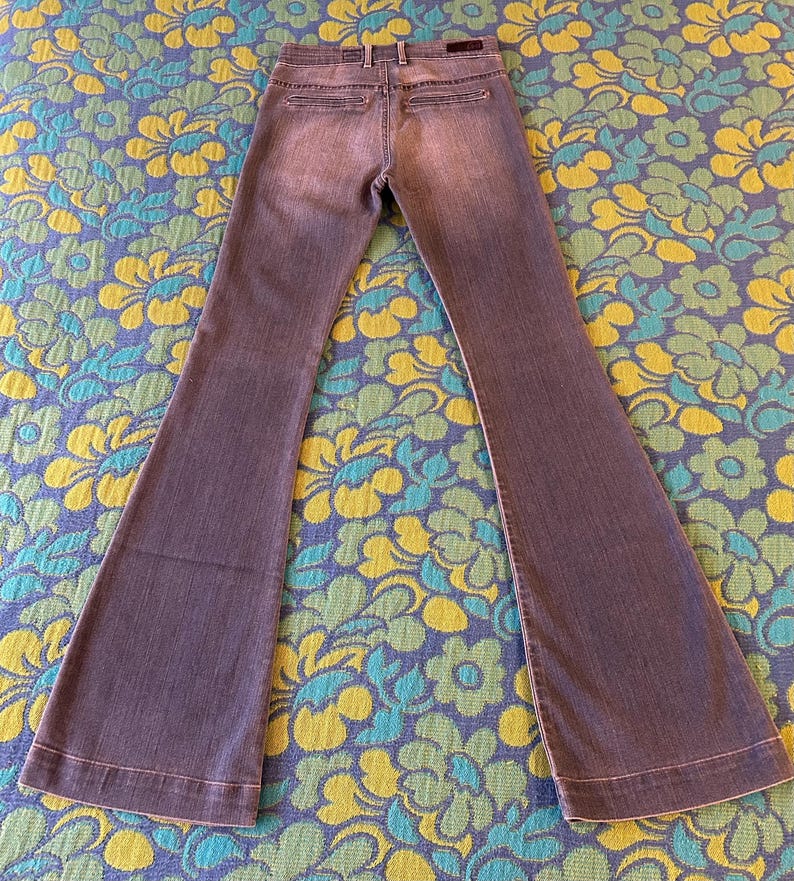 May include: A pair of gray denim bell-bottom jeans. The jeans have a classic five-pocket design, a button and zipper closure, and a flared leg. The jeans are laid flat on a floral patterned surface.