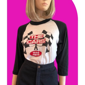 May include: A white and black baseball-style t-shirt with a black and white checkered flag design and the text "THE CARS 1980 TOUR" in red.