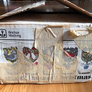 May include: A box of Anchor Hocking Christmas glasses with a white background and a red, green, and blue design. The box is labeled "Anchor Hocking" and "12 Days of Christmas".