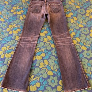 May include: Faded blue denim jeans with a flared leg design. The jeans have a worn appearance with lighter areas and detailed stitching on the back pockets. The jeans are laid flat on a floral patterned surface.