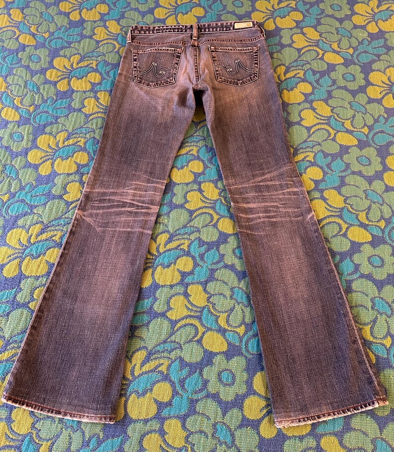May include: Faded blue denim jeans with a flared leg design. The jeans have a worn appearance with lighter areas and detailed stitching on the back pockets. The jeans are laid flat on a floral patterned surface.