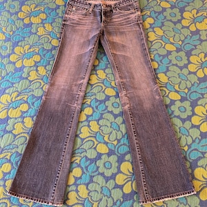 May include: Faded blue denim jeans with a flared leg design. The jeans feature a classic five-pocket style, a button and zip closure, and a worn appearance. The jeans are laid flat on a floral patterned surface.