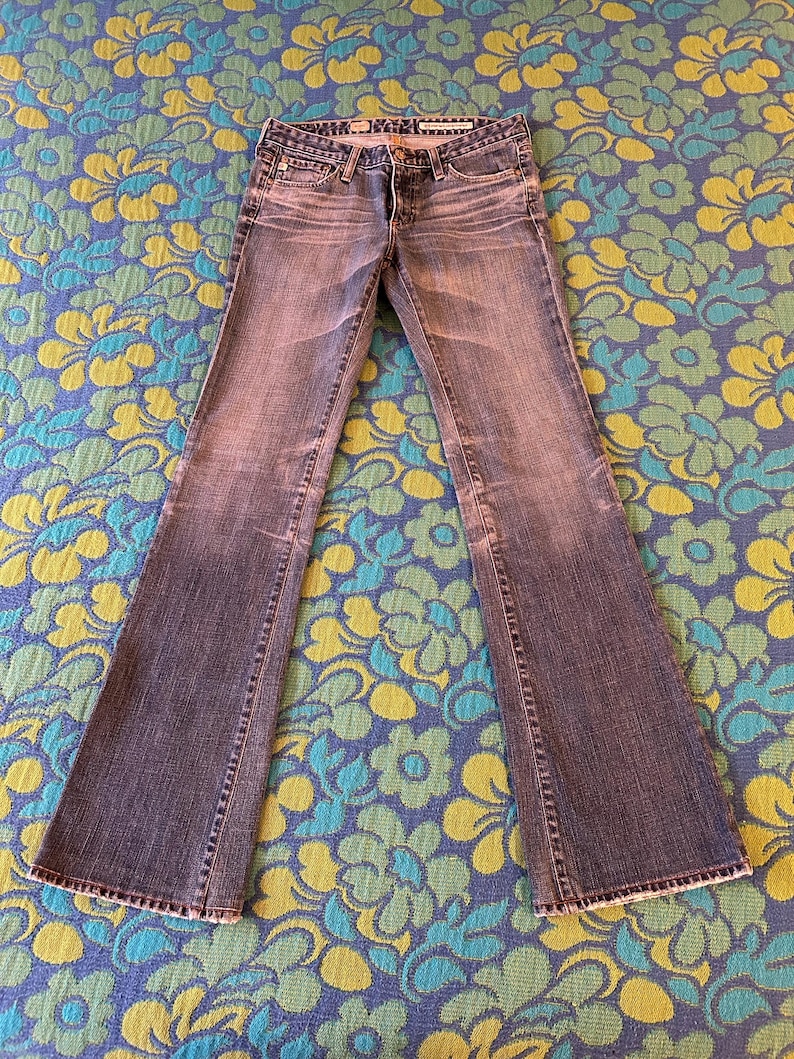 May include: Faded blue denim jeans with a flared leg design. The jeans feature a classic five-pocket style, a button and zip closure, and a worn appearance. The jeans are laid flat on a floral patterned surface.