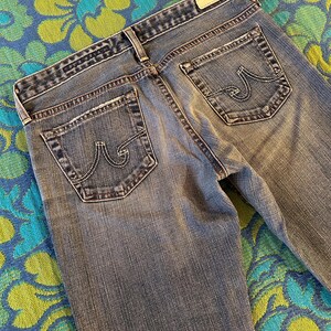 May include: Faded blue denim jeans with a classic five-pocket design. The jeans have a light wash, visible stitching, and the brand's logo on the back pockets. The jeans are laid flat, showing the back pockets and waistband.