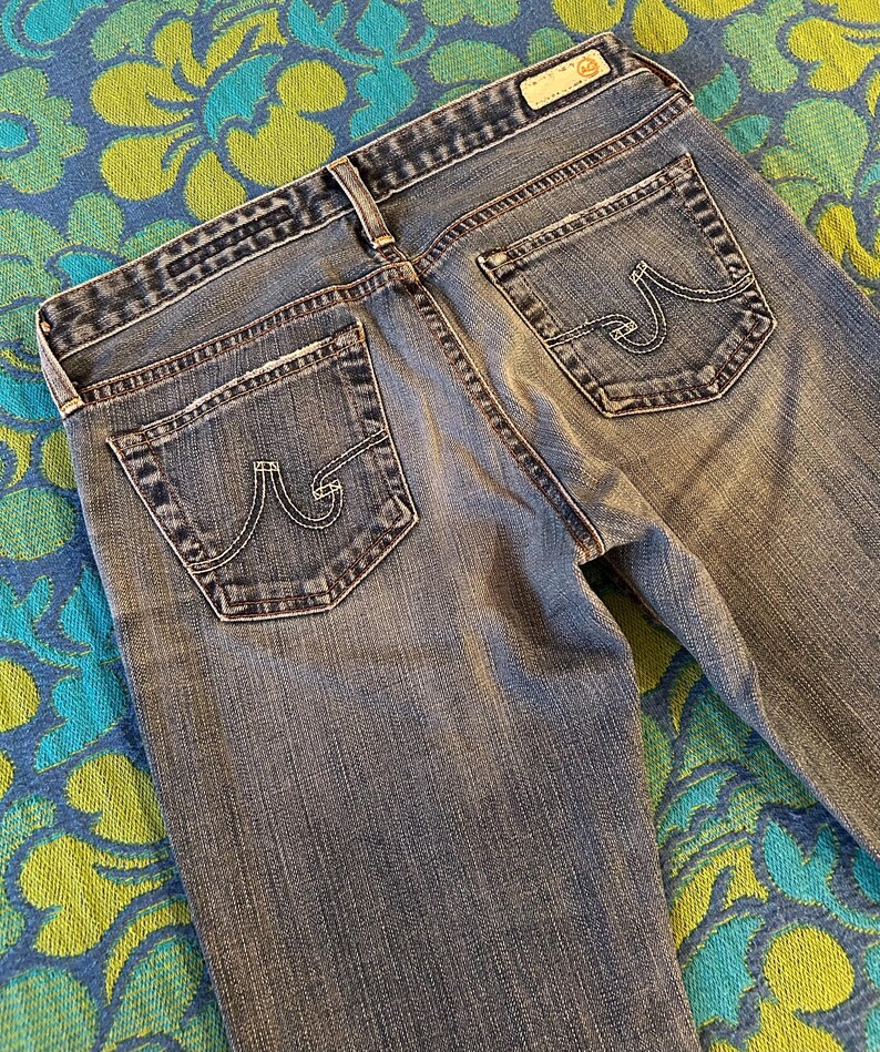 May include: Faded blue denim jeans with a classic five-pocket design. The jeans have a light wash, visible stitching, and the brand's logo on the back pockets. The jeans are laid flat, showing the back pockets and waistband.