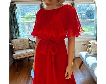 May include: A vibrant red dress with a pleated skirt and a matching tie belt. The dress features a flowing, cape-like top and is displayed on a mannequin. The dress is knee-length and has a ruffled hem.