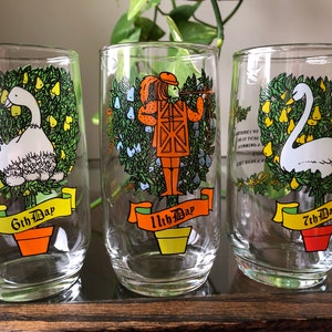 May include: Set of four clear glass tumblers with colorful illustrations of the 12 Days of Christmas. Each tumbler features a different day, including a swan, a pear tree, and a drummer boy. The tumblers have a yellow banner with the day number and the words "Day" printed on them.