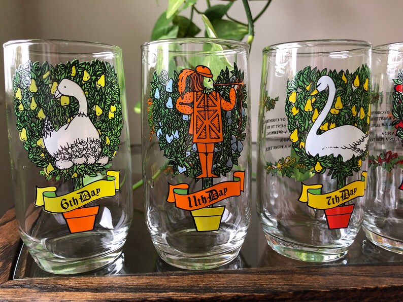 May include: Set of four clear glass tumblers with colorful illustrations of the 12 Days of Christmas. Each tumbler features a different day, including a swan, a pear tree, and a drummer boy. The tumblers have a yellow banner with the day number and the words "Day" printed on them.