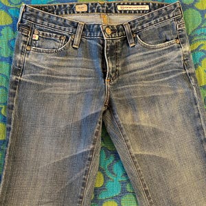 May include: Close-up of light blue denim jeans. The jeans have a classic five-pocket design, a button and zip closure, and belt loops. The jeans are slightly faded, with subtle wear marks.
