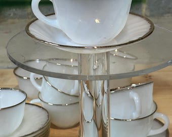 May include: A set of white milk glass teacups and saucers with a gold trim. The cups and saucers are stacked on a clear acrylic stand.