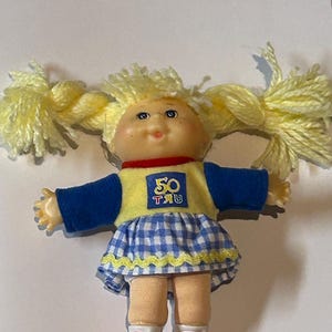May include: A small Cabbage Patch Kids doll with blonde pigtails wearing a blue and yellow outfit with a yellow patch that says "50 TRU".
