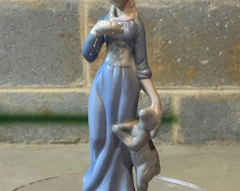 May include: A porcelain figurine of a woman in a blue dress holding a small lamb. The figurine is standing on a base with a rocky texture.