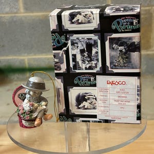 May include: A porcelain figurine ornament of a girl wearing a brown hat and a white dress holding a bouquet of flowers. The ornament is sitting on a small box with the words "You bring a smile to everyone" printed on it. The ornament is part of the "Kim Anderson's As A Picture" collection by Enesco.