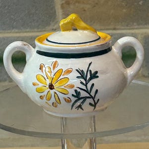 May include: A white ceramic sugar bowl with a yellow lid and floral design. The bowl has two handles and a yellow flower with green leaves painted on the side.