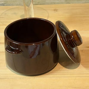 May include: A brown ceramic crock pot with a lid. The crock pot has two handles on the side and a white rim. The lid has a brown knob.