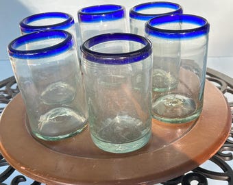 May include: Six clear glass tumblers with a vibrant blue rim, arranged on a round, copper-toned tray. The glasses are cylindrical and appear to be hand-blown. The tray sits on a decorative black metal table, creating a striking contrast.
