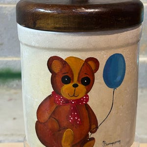 May include: A ceramic cookie jar with a wooden lid. The jar is decorated with a hand-painted image of a brown teddy bear wearing a red and white polka dot scarf and holding a blue balloon. The artist's signature, "N.J. Hoffmaster", is visible on the bottom of the jar.