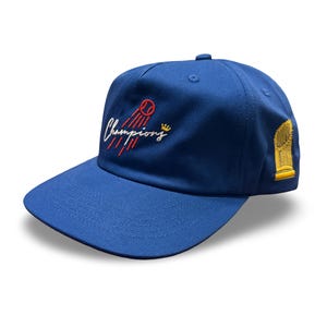 Op de afbeelding: Koningsblauwe baseballcap met geborduurde tekst "Champions" en een trofee-afbeelding. De cap heeft een gebogen klep en een gestructureerde kroon. De tekst en afbeeldingen zijn in wit, rood en goud.