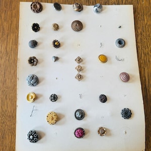 Könnte beinhalten: Eine Sammlung von Vintage-Knöpfen in verschiedenen Formen, Größen und Farben, darunter Blumen-, Wirbel- und geometrische Designs. Die Knöpfe sind auf einer cremefarbenen Karte angeordnet, die ihre komplizierten Details und Texturen hervorhebt. Einige Knöpfe haben ein metallisches Finish.