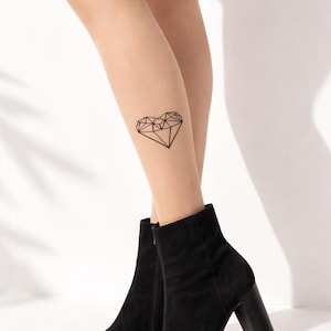 Op de afbeelding: Zwarte suède enkellaarsjes met blokhak. Een geometrische harttattoo is zichtbaar op het been. De laarzen hebben een spitse neus en een zijrits. De achtergrond is wit.