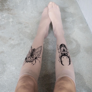 May include: Nude-coloured sheer tights with black insect illustrations. One leg displays a cicada design, the other a beetle. The tights are worn against a grey background.
