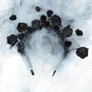 May include: A black floral headband with fabric roses and buds. The headband is set against a backdrop of white and gray tulle fabric. The flowers are clustered on the headband, with some extending beyond the band.
