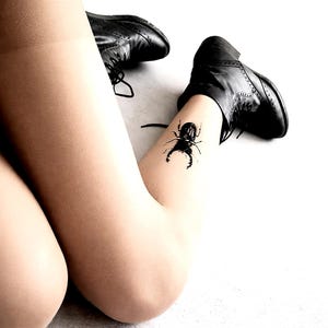 May include: Black leather lace-up boots and a black beetle tattoo on a leg. The legs are covered in sheer, light-colored hosiery. The boots are on a white surface.
