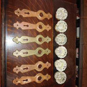 May include: Six brass door plates with keyholes and circular openings arranged in a column, paired with six clear, faceted glass door knobs. The metal plates feature a decorative, elongated shape with pointed ends and screw holes.